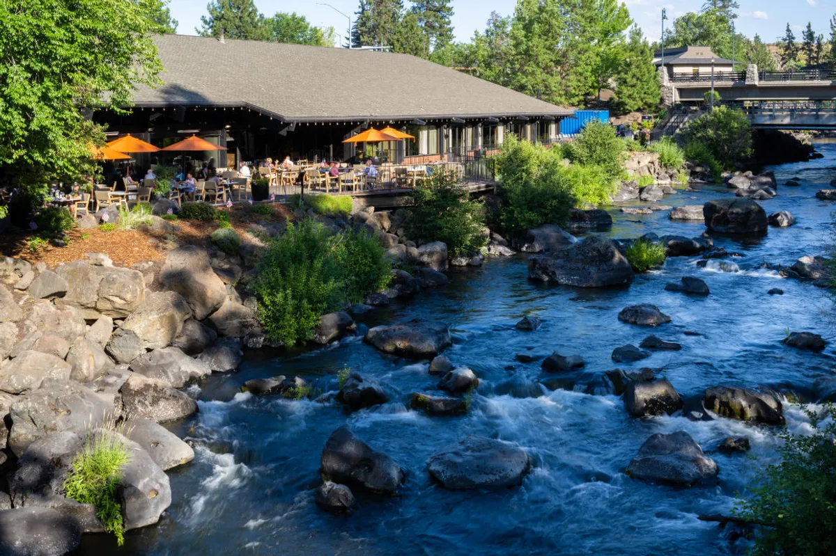 The image showcases a vibrant restaurant patio featuring orange umbrellas set alongside the flowing Deschutes River. The rocky shoreline and lush greenery frame the scene, creating a tranquil and inviting atmosphere perfect for outdoor dining, social gatherings, and relaxing by the water.