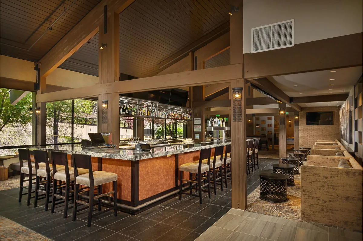 A spacious, modern lounge and bar area filled with natural light from oversized windows. The bar counter is well-stocked, with comfortable seating areas arranged for casual gatherings, and the vibe is lively and welcoming.