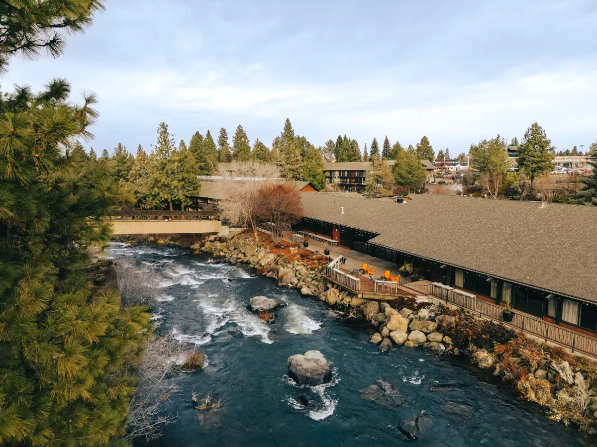 This peaceful exterior shot features a deck area overlooking the river, with the building partially visible, surrounded by trees. The tranquil setting highlights both the natural beauty and the inviting outdoor space, reminiscent of a private balcony or patio experience.