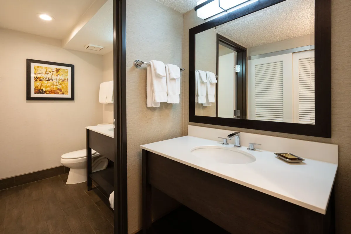 This image shows a bathroom vanity with a sink, framed artwork above, and plush towels neatly arranged on a rack. The space is bright and inviting, with crisp, clean surfaces and a sense of order that evokes a hotel-quality environment.