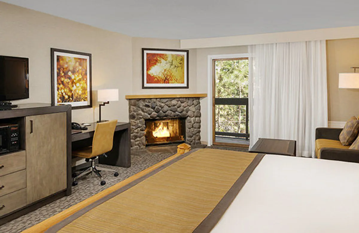 This interior shot highlights a stylish studio corner with a modern fireplace, a bed, striking artwork, and a window. The combination of workspace, cozy fire, and tranquil setting captures the adaptability of the room for both work and relaxation.