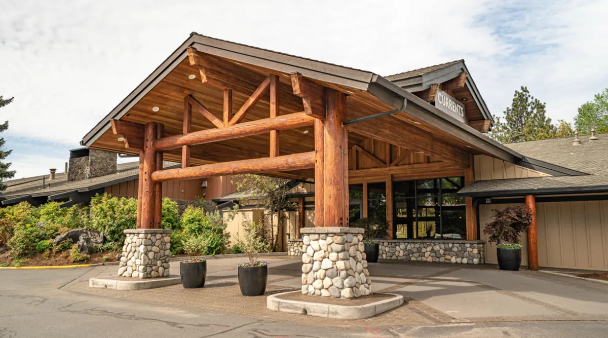 A welcoming exterior with massive wooden beams, stone columns, and a glass entrance, set against a warm evening backdrop. People are gathered on the patio, enjoying meals and events as the sun sets.