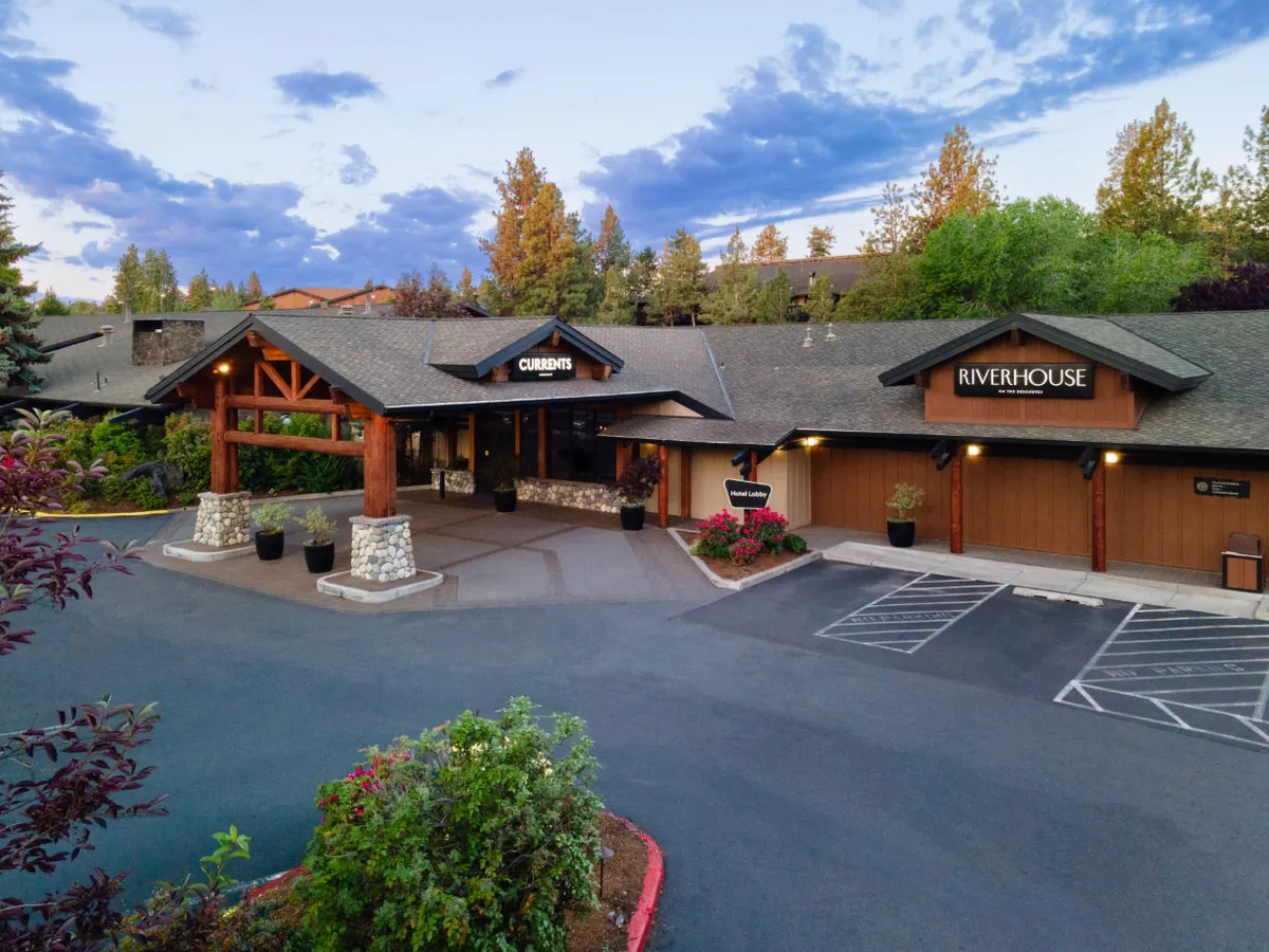 A vibrant exterior view highlighting the signage for the CURRENTS Lounge and RiverHouse entrances, surrounded by manicured landscaping. The scene captures groups enjoying sunset dining on the patio and the courtyard set up for community celebrations and special occasions.