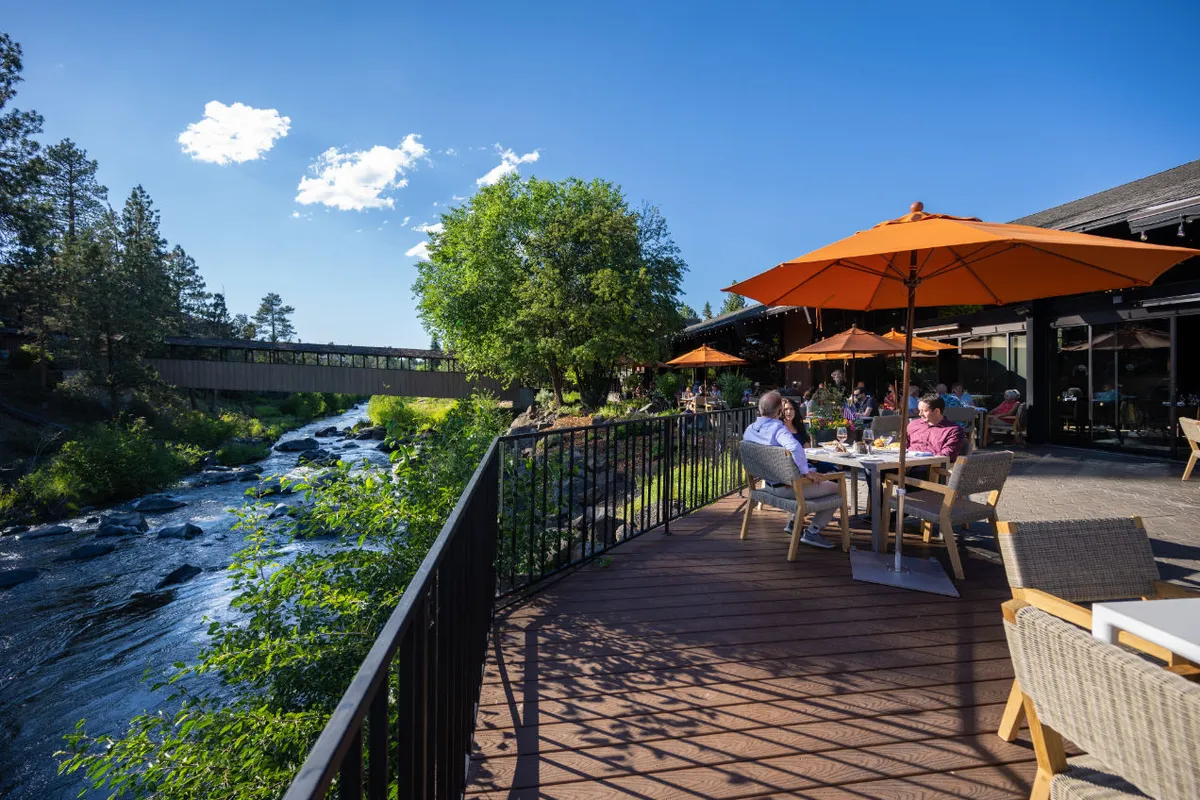 This exterior image showcases a scenic riverfront setting with outdoor seating areas framed by orange umbrellas, lush trees, and the gently flowing river. The arrangement evokes a sense of relaxation and immersion in nature, with the seating positioned to enjoy direct views of the water and surrounding landscape.
