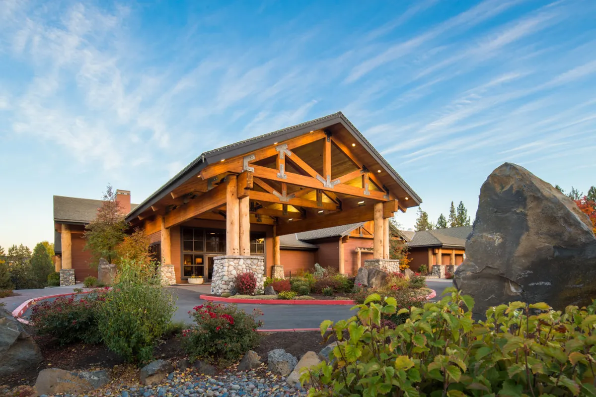 An inviting exterior view with large wooden beams, a striking stone facade, and landscaped gardens with rock formations. Guests are seen gathering outdoors, enjoying the peaceful natural surroundings.