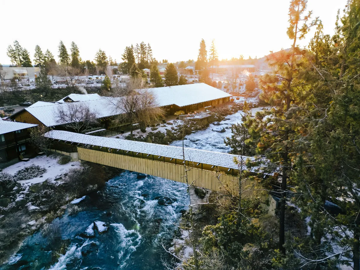 A tranquil riverside scene featuring a covered bridge spanning a flowing river, surrounded by lush trees and snow-dusted rooftops. People are seen walking across the bridge, taking in the beauty of the sunset and the natural setting.
