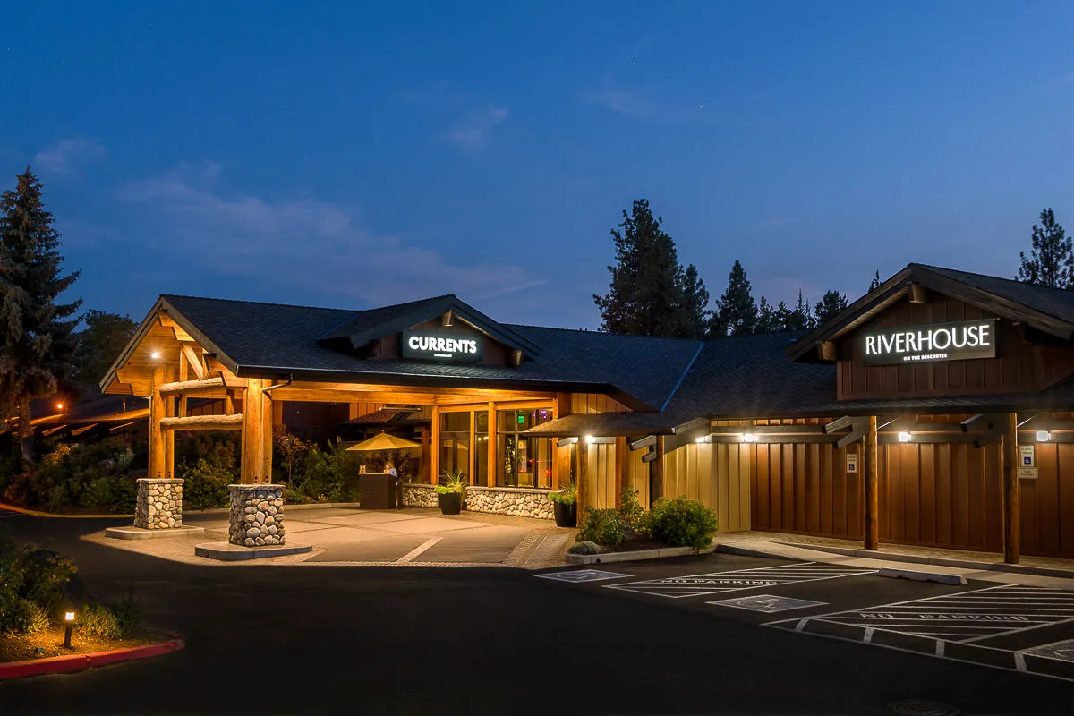 This exterior image showcases the main entrance framed by warm lighting fixtures, a striking wooden structure, and lush landscape greenery. Diners are visible enjoying intimate meals under the stars, while small groups gather in a cozy, welcoming atmosphere.